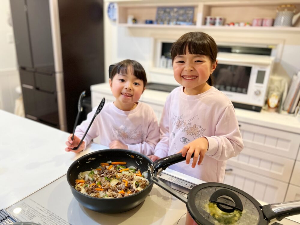 野菜炒めの入ったフライパンを前に微笑む姉妹の子どもたち