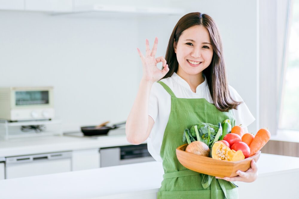 野菜のはいった籠を持ちながら、手で丸印を作る女性の写真